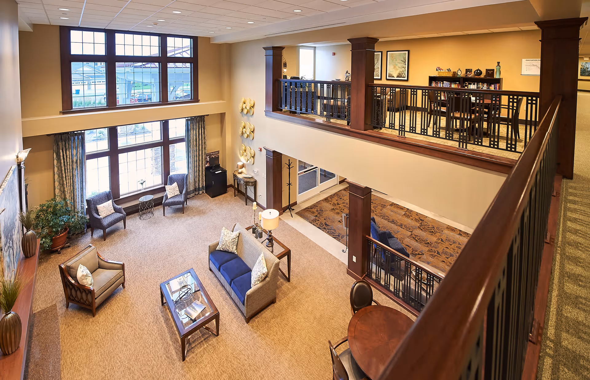 Spacious two-story living room area with large windows letting in natural light. The lower level features a blue sofa, armchairs, a coffee table, and potted plants. The upper level has a railing with a high table and chairs, along with framed artwork on the walls. The decor includes warm tones and wood accents.