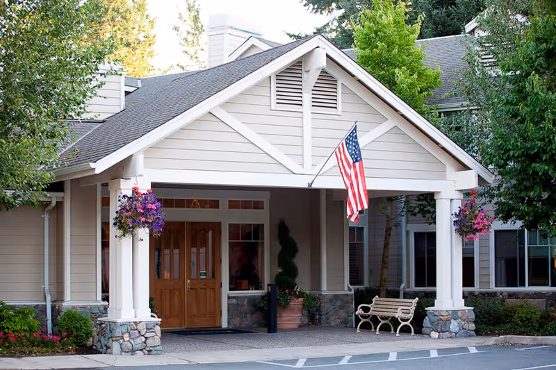 Entrance of a senior living facility with a covered porch supported by white columns and stone bases. There are hanging flower baskets on both sides of the porch, an American flag mounted on the right column, a wooden bench, and a double wooden door entrance. The building exterior is beige with stone accents and surrounded by trees and shrubs.