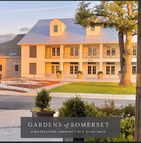 Exterior view of a two-story building with a metal roof and multiple windows, surrounded by greenery and a paved driveway. The building has a porch with columns and potted plants, and the address number 340 is visible above the entrance.