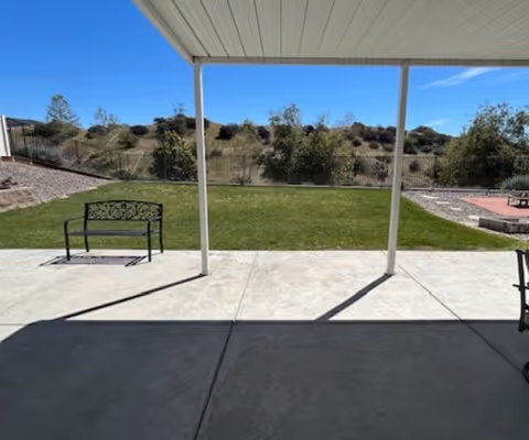 View from a covered patio looking out onto a grassy yard with a metal bench on the left side. There are trees and shrubs beyond a fence, with hills in the background under a clear blue sky.