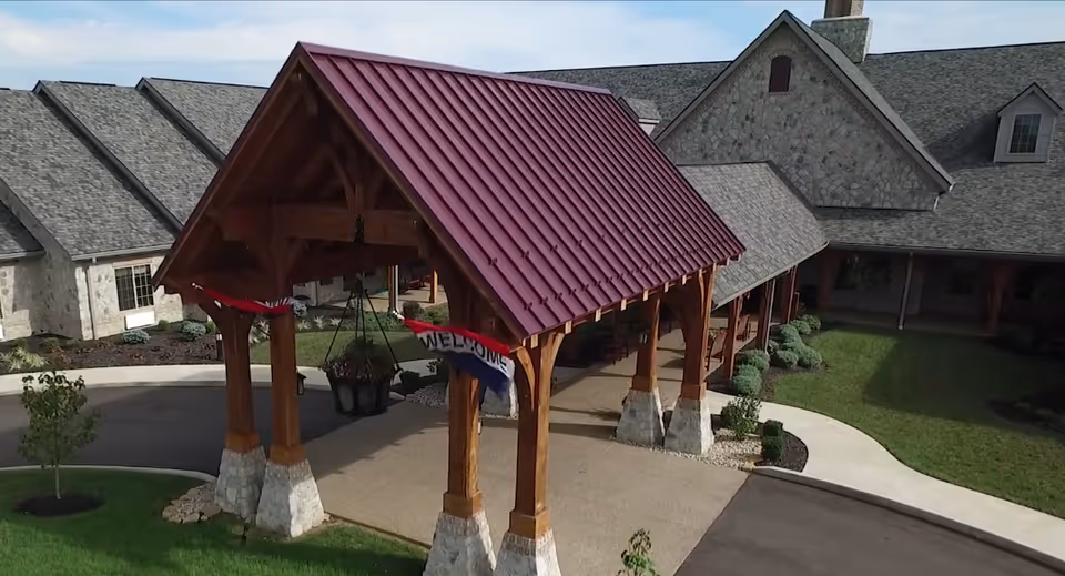 Entrance of The Inn at Winchester Trail featuring a wooden porte-cochère with a red metal roof and stone bases on the wooden pillars. The building has stone walls and gray shingled roofs. A red, white, and blue welcome banner hangs from the structure. There is a driveway and landscaped greenery around the entrance.