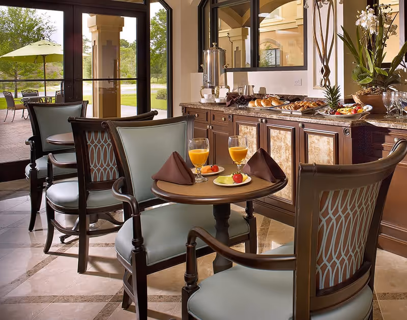 Small dining area with round tables and cushioned chairs set with glasses of orange juice and plates of fruit beside a buffet and glass doors opening to an outdoor patio.