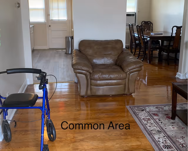 A common area in an assisted living facility featuring a brown leather armchair in the center, a blue walker on the left, a wooden dining table with chairs in the background, hardwood floors, and a rug partially visible on the right.