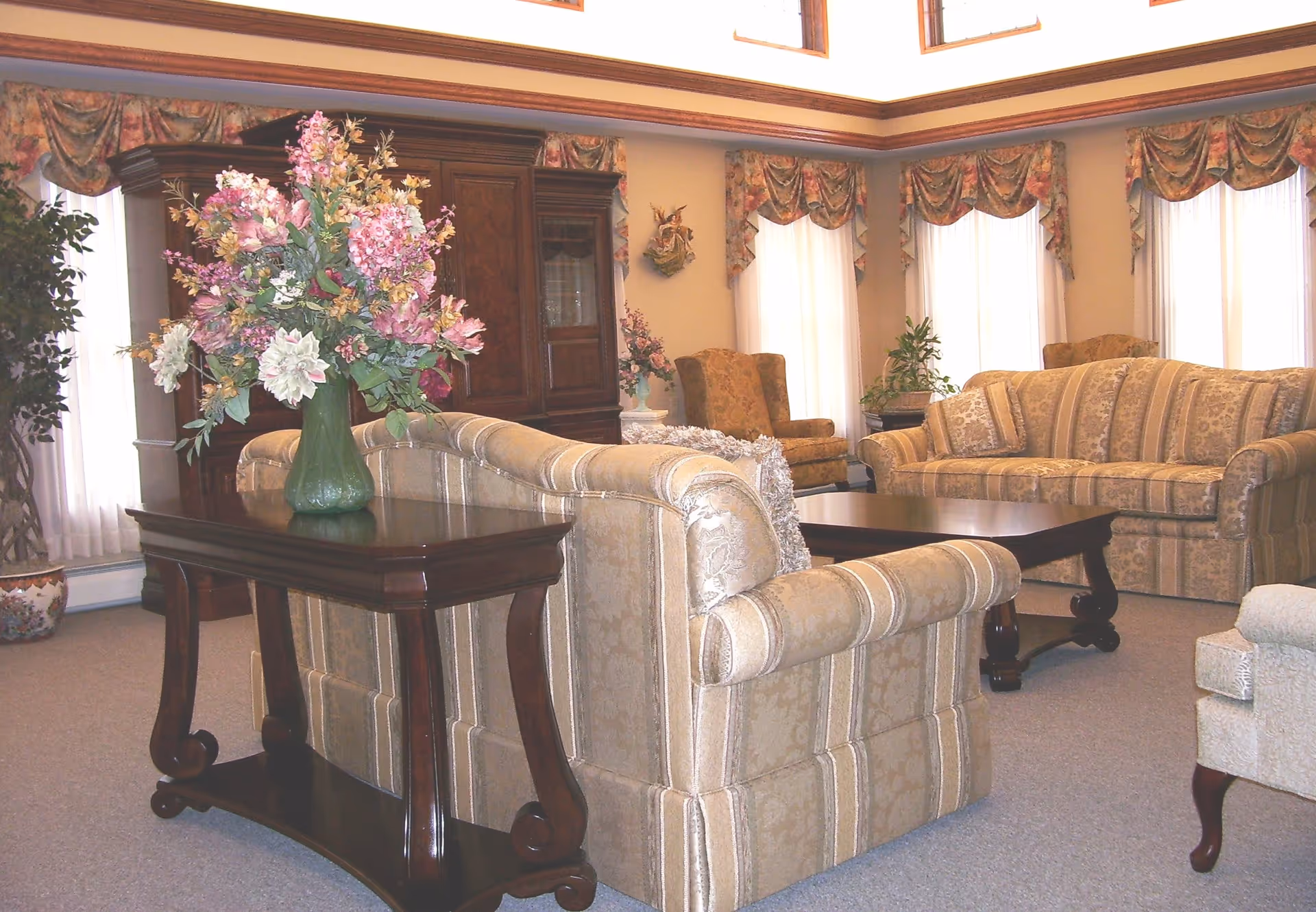 A spacious living room with beige patterned sofas and armchairs arranged around a dark wooden coffee table. A dark wooden console table behind one sofa holds a large vase with pink and white flowers. The room has large windows with sheer white curtains and floral valances, and a tall wooden cabinet against the wall.