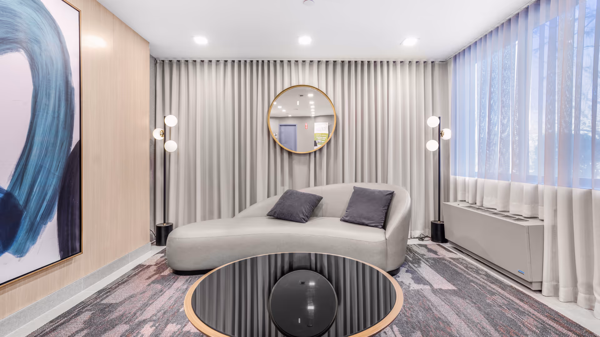 Modern lounge area with a curved light gray sofa with two dark gray pillows, a round black glass coffee table with a gold rim, two tall floor lamps with three white globe lights each, a large abstract painting on the left wall, a round mirror on the back wall with gray curtains, and a large window with sheer white curtains on the right side.