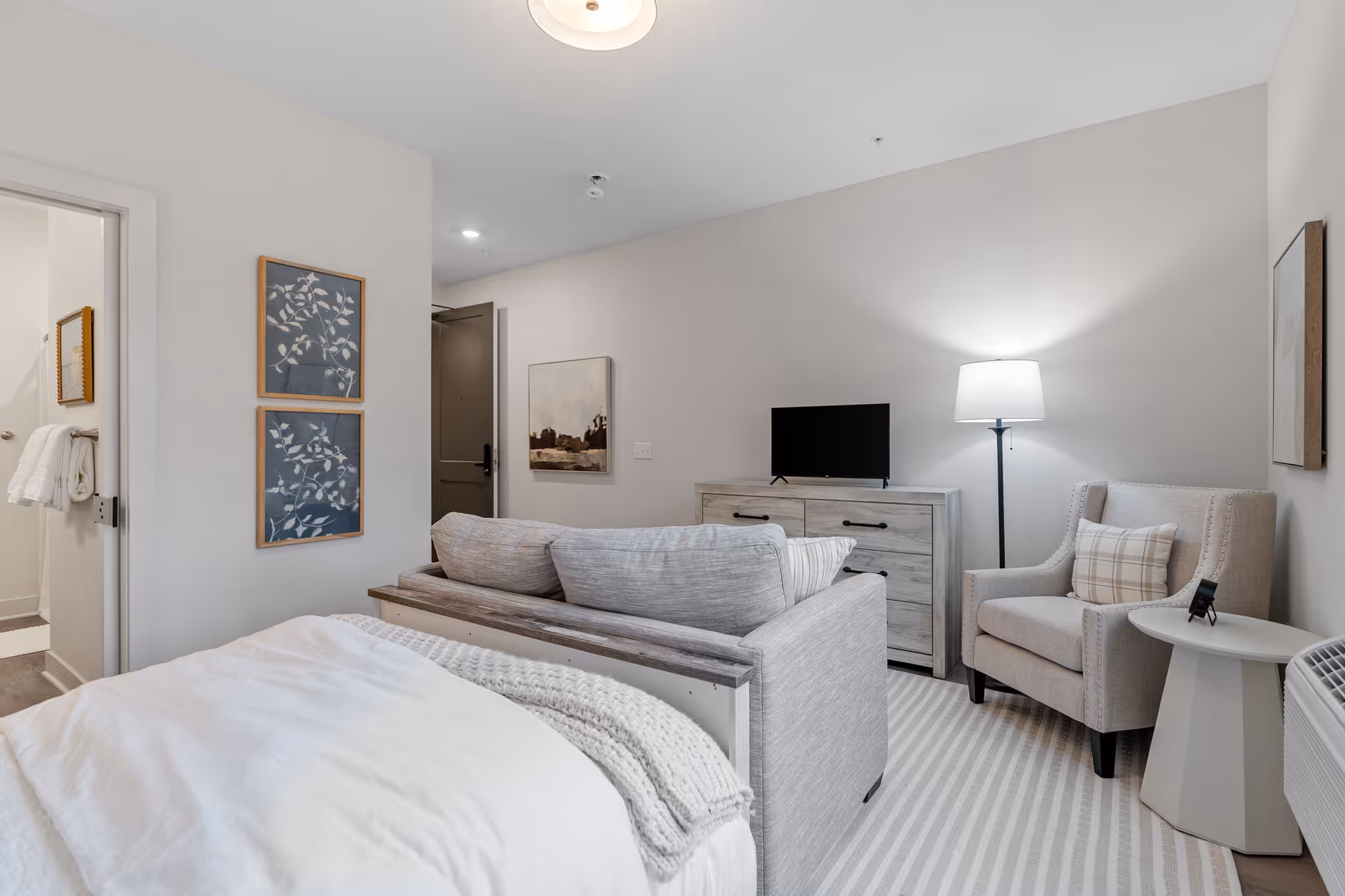 Neutral-toned bedroom featuring a bed, sofa at the foot of the bed, dresser with a small TV, and a reading chair with a floor lamp.