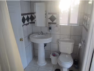 Small bathroom with white tiled walls featuring a diamond pattern border, a pedestal sink with a mirror cabinet above it, a toilet, a small trash bin, and a window letting in sunlight.