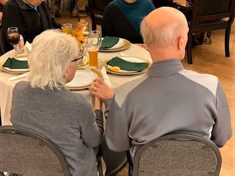 Two elderly people sitting side by side at a dining table in a senior living facility, holding hands. The table is set with plates, green napkins, glasses, and a small centerpiece.