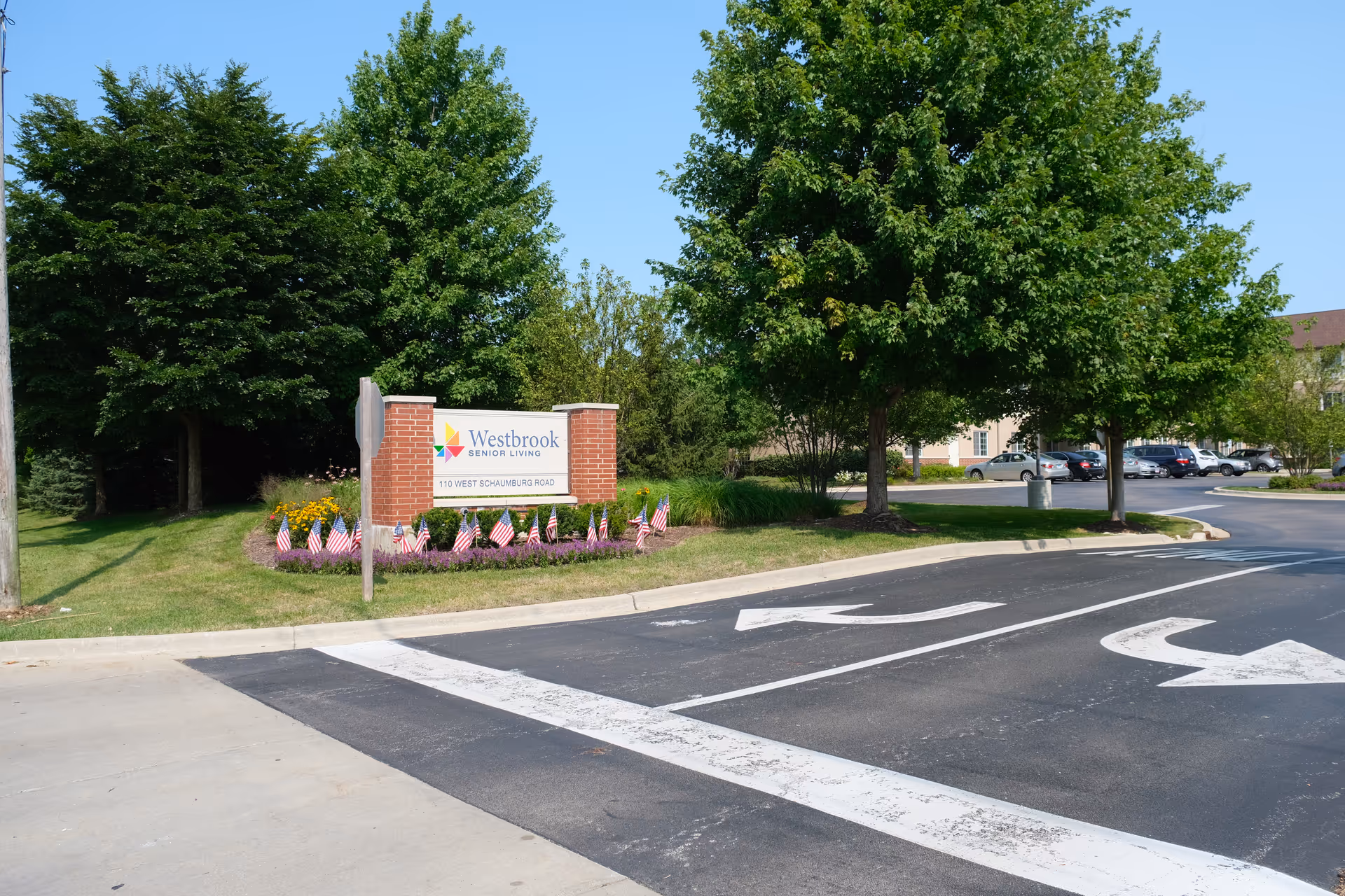 Entrance sign for Westbrook Senior Living at 110 West Schaumburg Road, surrounded by green trees, small American flags, and a parking lot with several cars in the background under a clear blue sky.