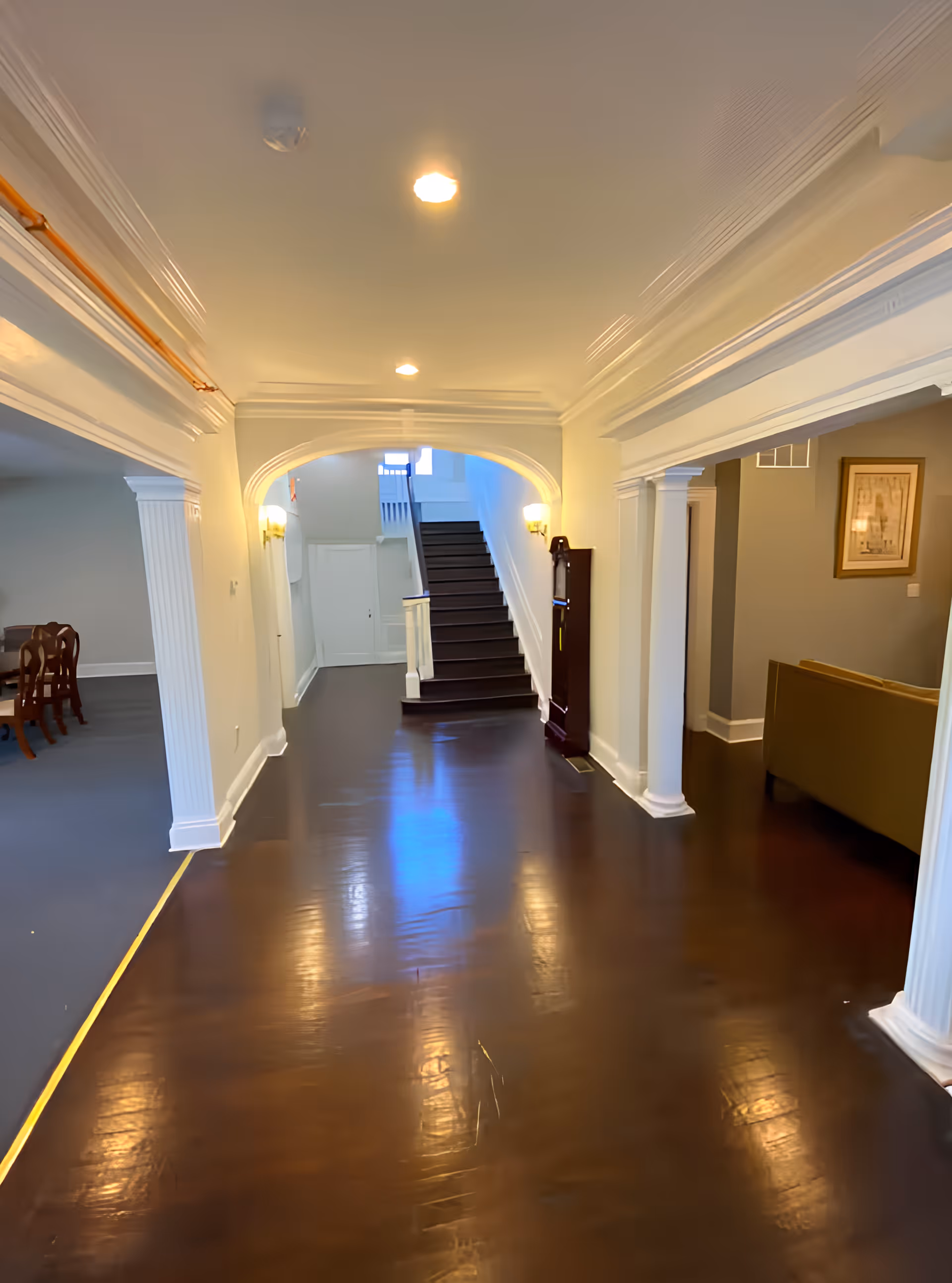 Interior hallway of an assisted living facility with polished dark wooden floors, white walls, and ceiling lights. The hallway leads to a staircase at the end, with a grandfather clock on the right side. On the left, there is a dining area with a wooden dining table and chairs, and on the right, a living area with a couch and framed artwork on the wall.