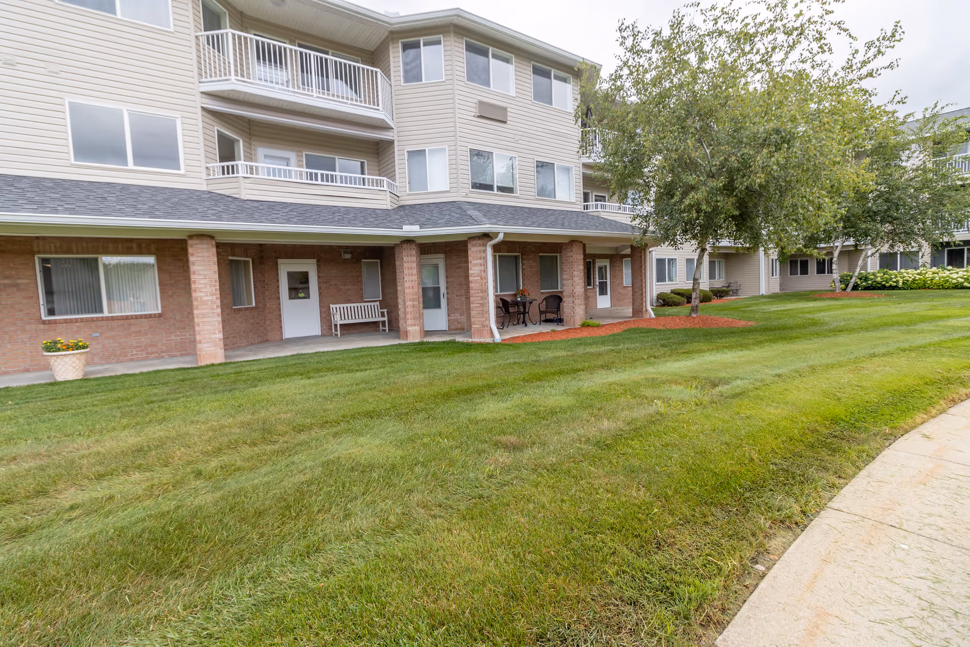 Exterior view of a multi-story senior living facility with beige siding and brick accents. The building features balconies, a covered patio area with seating, well-maintained green lawn, trees, and a curved concrete walkway.