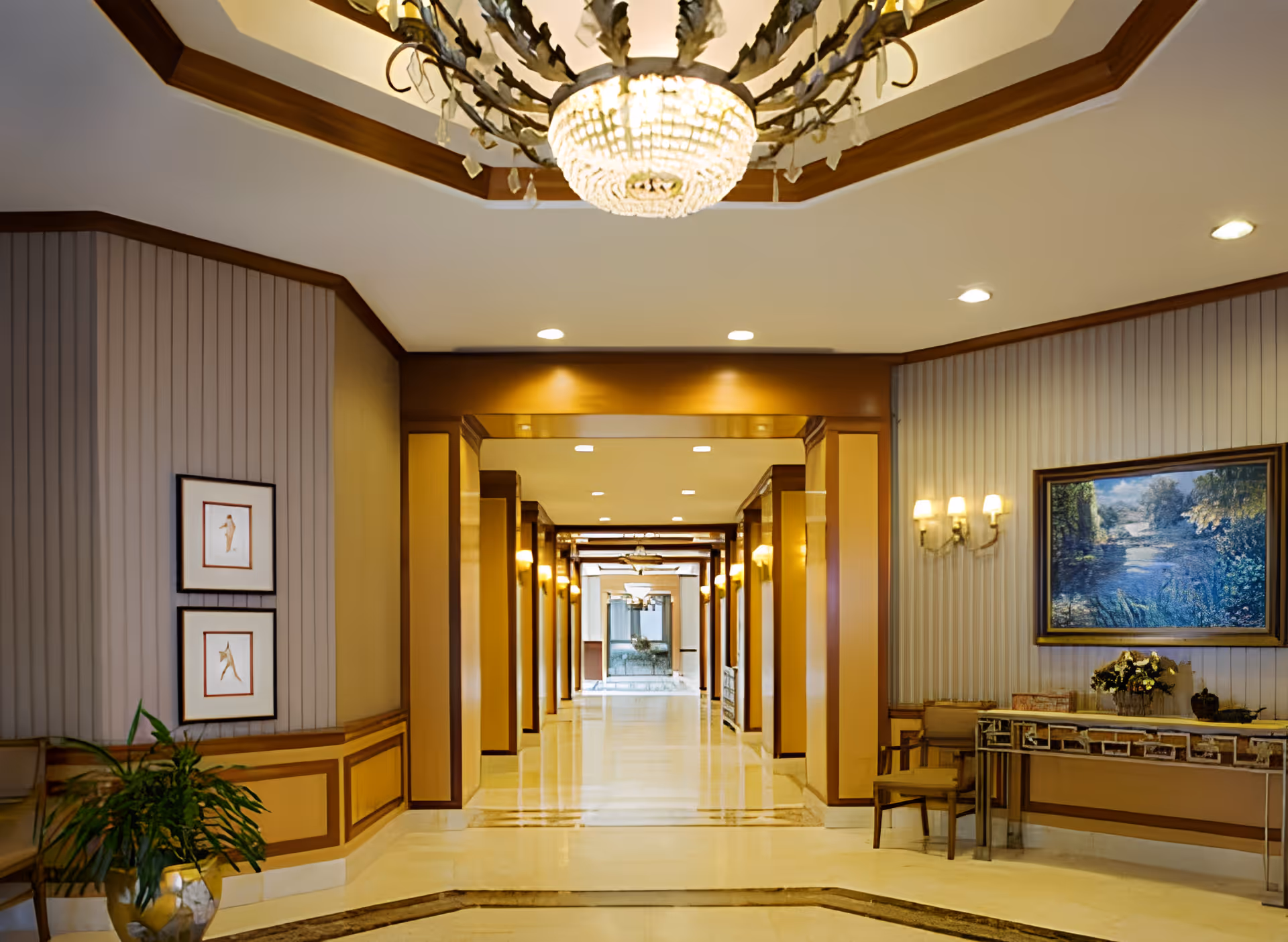 A well-lit, elegant hallway in a senior living facility with polished floors, decorative wall paneling, framed artwork, a chandelier, and a console table with chairs and a flower arrangement.