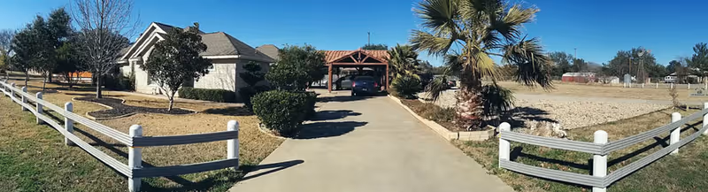 A driveway leading to a house with a car parked under a wooden carport, surrounded by a white fence, palm trees, and other greenery under a clear blue sky.