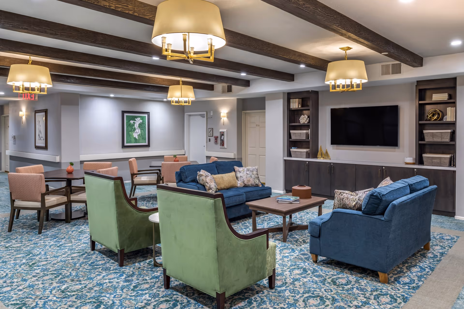 A spacious and well-lit common area with blue sofas, green armchairs, and several tables with pink chairs. The room features a patterned blue carpet, wooden ceiling beams, and modern pendant lights. A large flat-screen TV is mounted on the wall above built-in dark wood cabinets and shelves with decorative items and books. Artwork is hung on the walls, and an exit sign is visible in the background.
