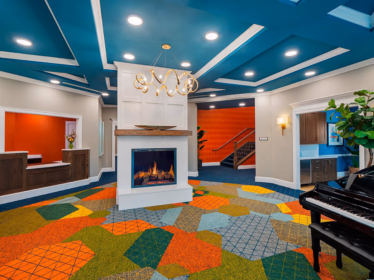 A brightly colored interior common area with a modern fireplace in the center, a blue coffered ceiling with recessed lighting, a geometric patterned carpet in orange, green, yellow, and blue tones, a black grand piano on the right, and a reception desk with a red-orange wall in the background.