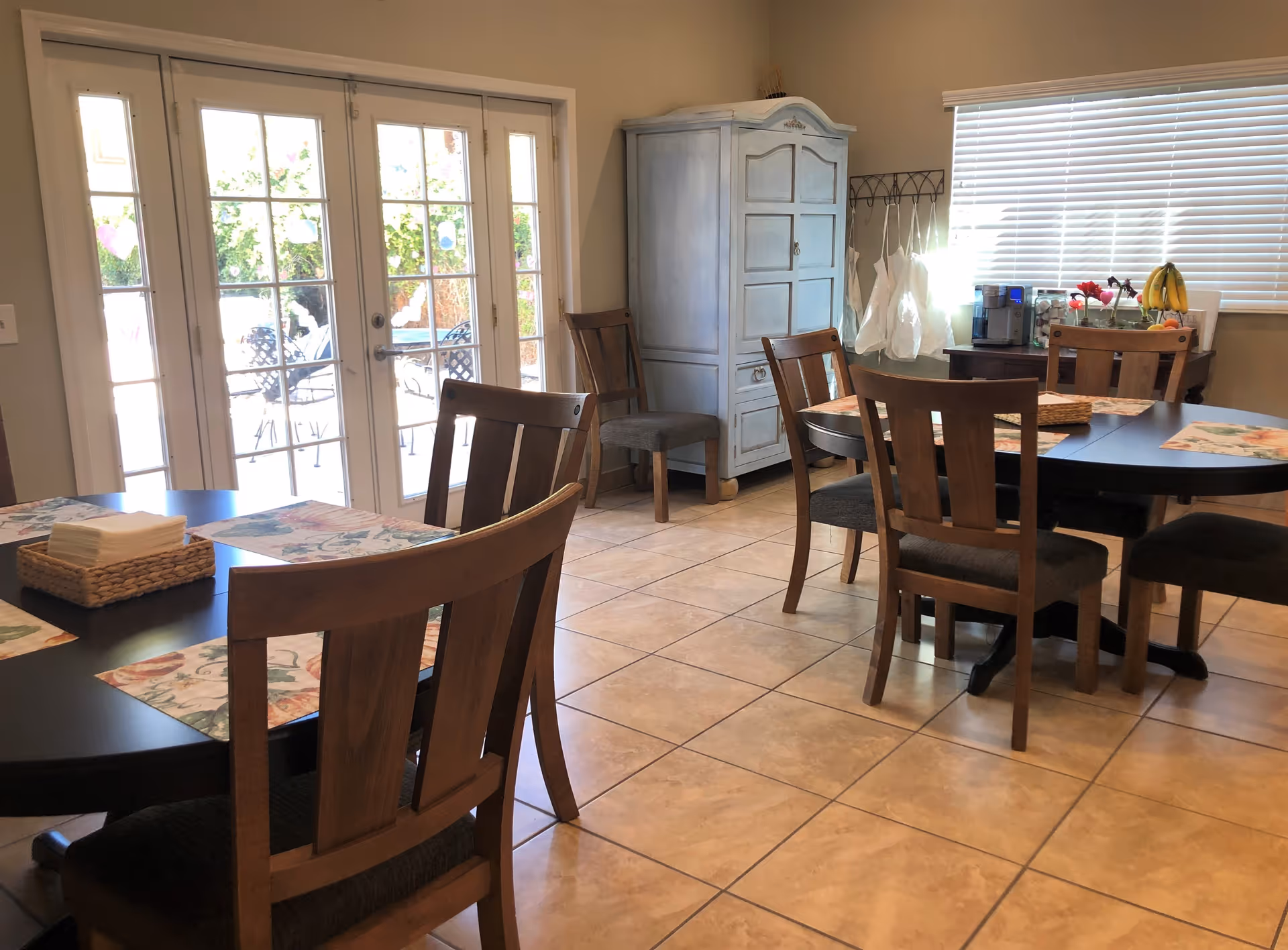 A dining area with two round black tables surrounded by wooden chairs with dark cushions. The tables have floral placemats and a basket with napkins. There is a light blue cabinet against the wall, a window with blinds, and a small table with a coffee machine, fruit bowl, and flowers. French doors lead outside to a patio with metal chairs and a table.