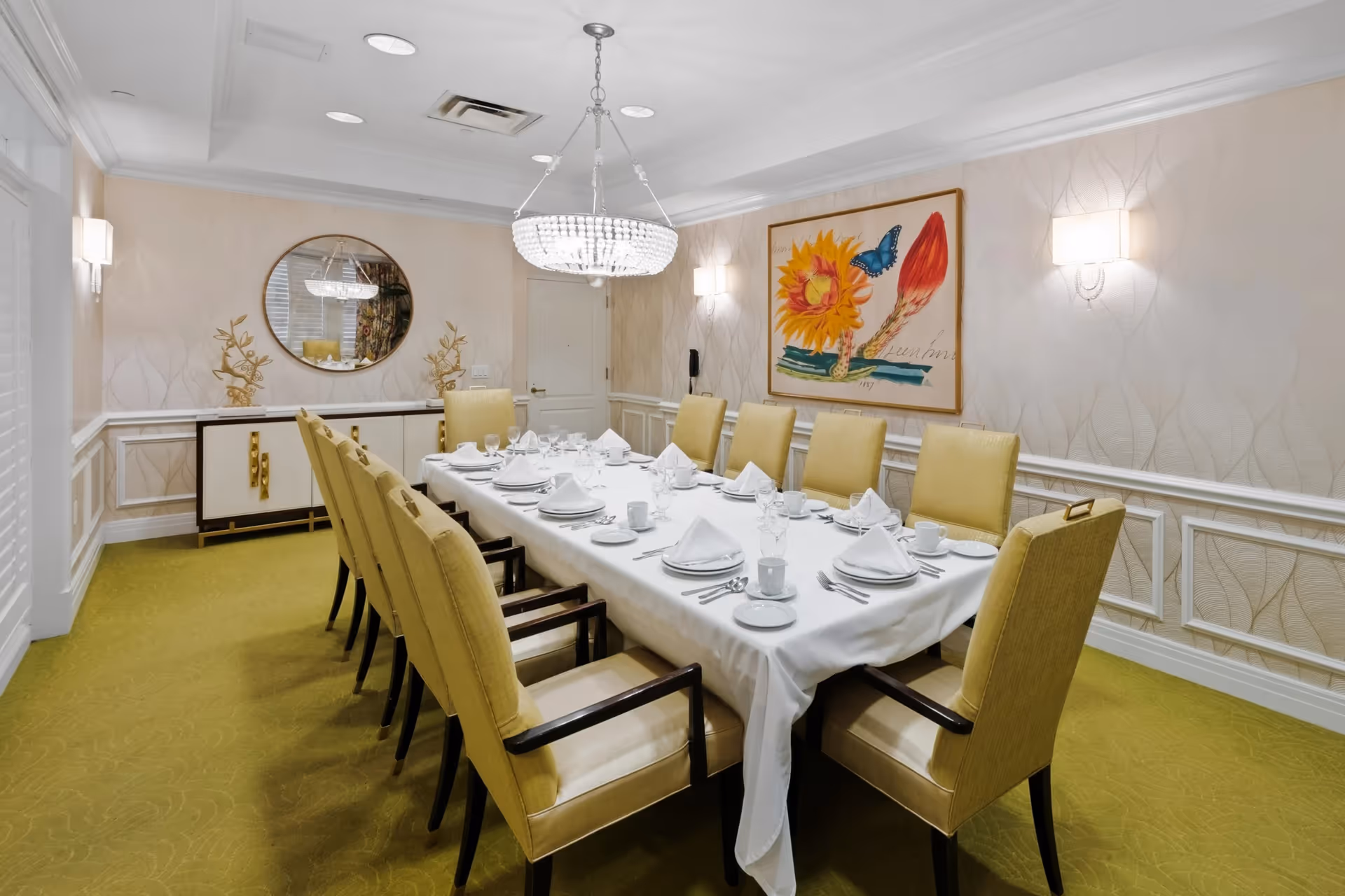 A formal dining room with a long rectangular table covered with a white tablecloth, set with white plates, cups, napkins, and silverware. The table is surrounded by ten yellow upholstered chairs with dark wooden arms and legs. The room features light-colored wallpaper with a subtle pattern, a green carpet, a modern chandelier hanging above the table, wall sconces, a round mirror, decorative gold sculptures on a sideboard, and a large framed artwork of a sunflower and a butterfly on the wall.