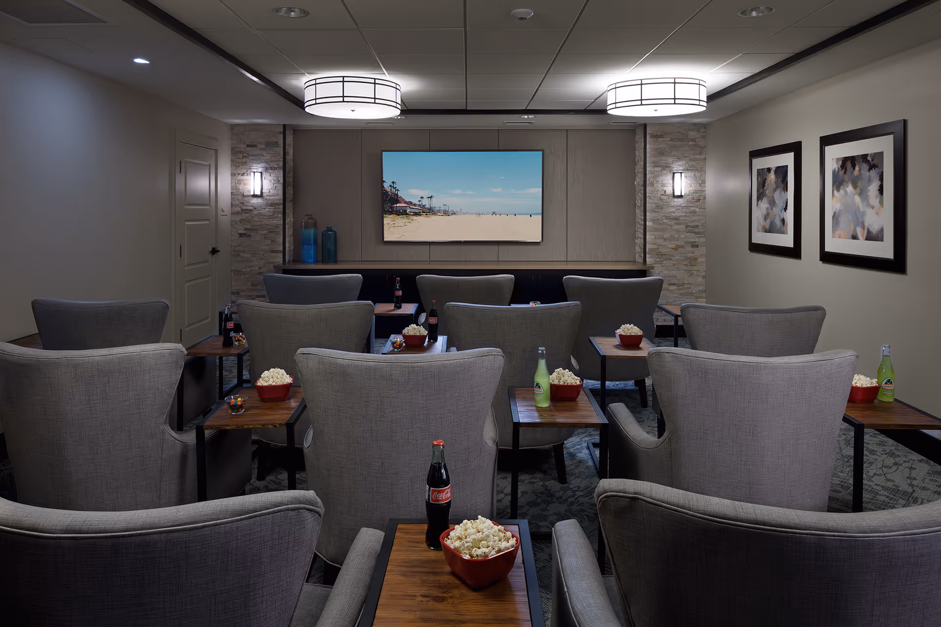 A small theater room with several rows of gray upholstered armchairs, each with a small wooden side table holding a bowl of popcorn and a bottle of soda. A large flat-screen TV is mounted on the front wall displaying a beach scene. The room has soft lighting with two ceiling lights and wall sconces, and two framed abstract paintings on the right wall.