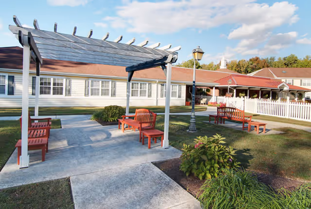 Outdoor seating area at Dover Place featuring a white pergola with red benches underneath, a concrete walkway, a vintage-style street lamp, green bushes, and a white picket fence surrounding the building with a red roof in the background under a partly cloudy sky.