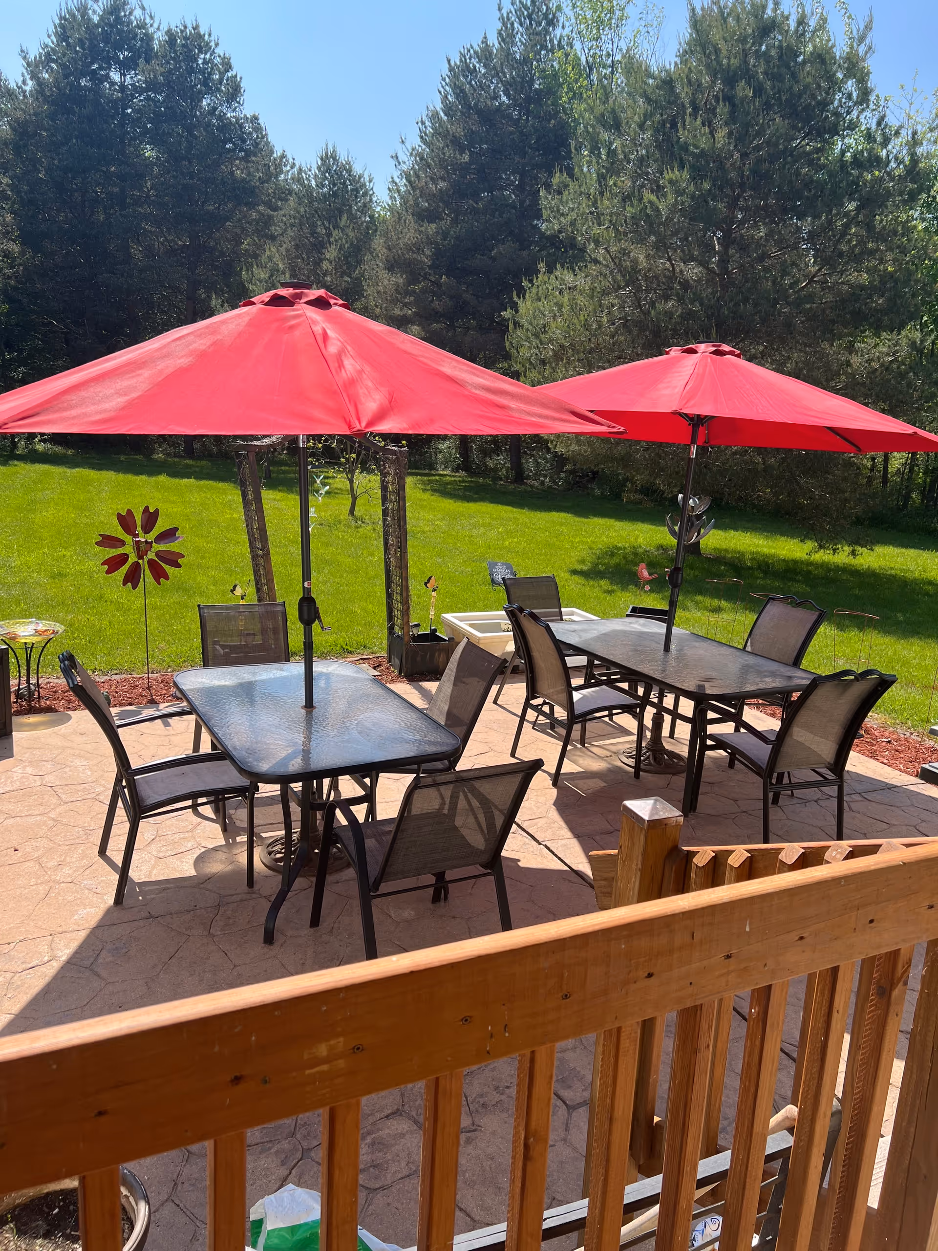 Outdoor patio area with two glass tables, each surrounded by four chairs. Both tables have large red umbrellas providing shade. The patio is paved and bordered by a wooden railing, with a green grassy area and trees in the background under a clear blue sky.