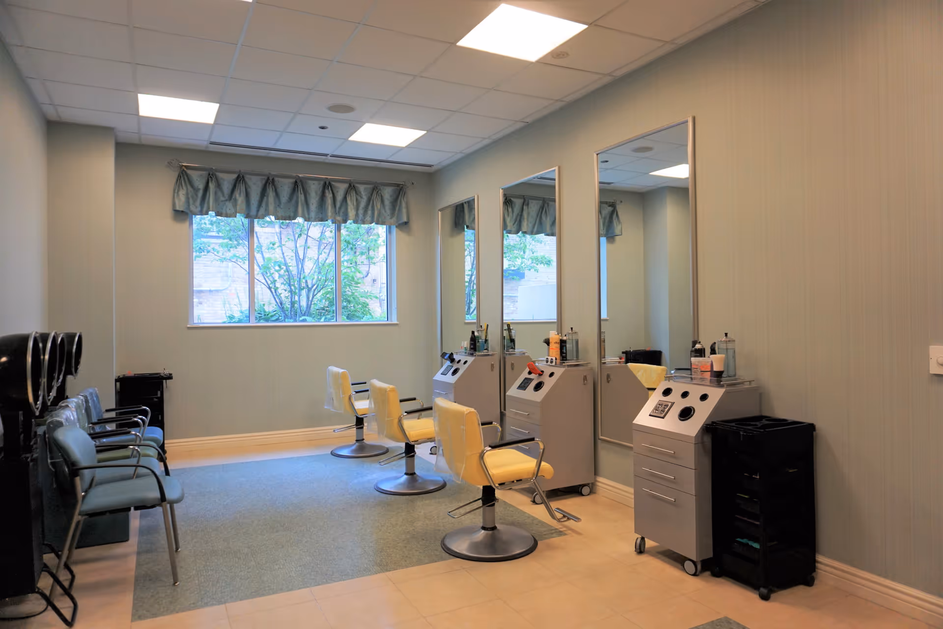Interior view of a salon area with two yellow salon chairs facing large mirrors mounted on the wall. There are hair styling stations with various hair products and tools. To the left, there are several blue waiting chairs and hair dryers. A window with a blue valance lets in natural light.
