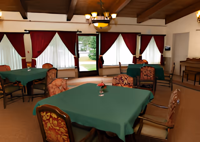 Interior view of a dining room with several tables covered in green tablecloths and surrounded by upholstered chairs. The room has large windows with red curtains and white sheer drapes, a wooden ceiling with exposed beams, and a chandelier light fixture. There is a piano against one wall and a door leading outside.