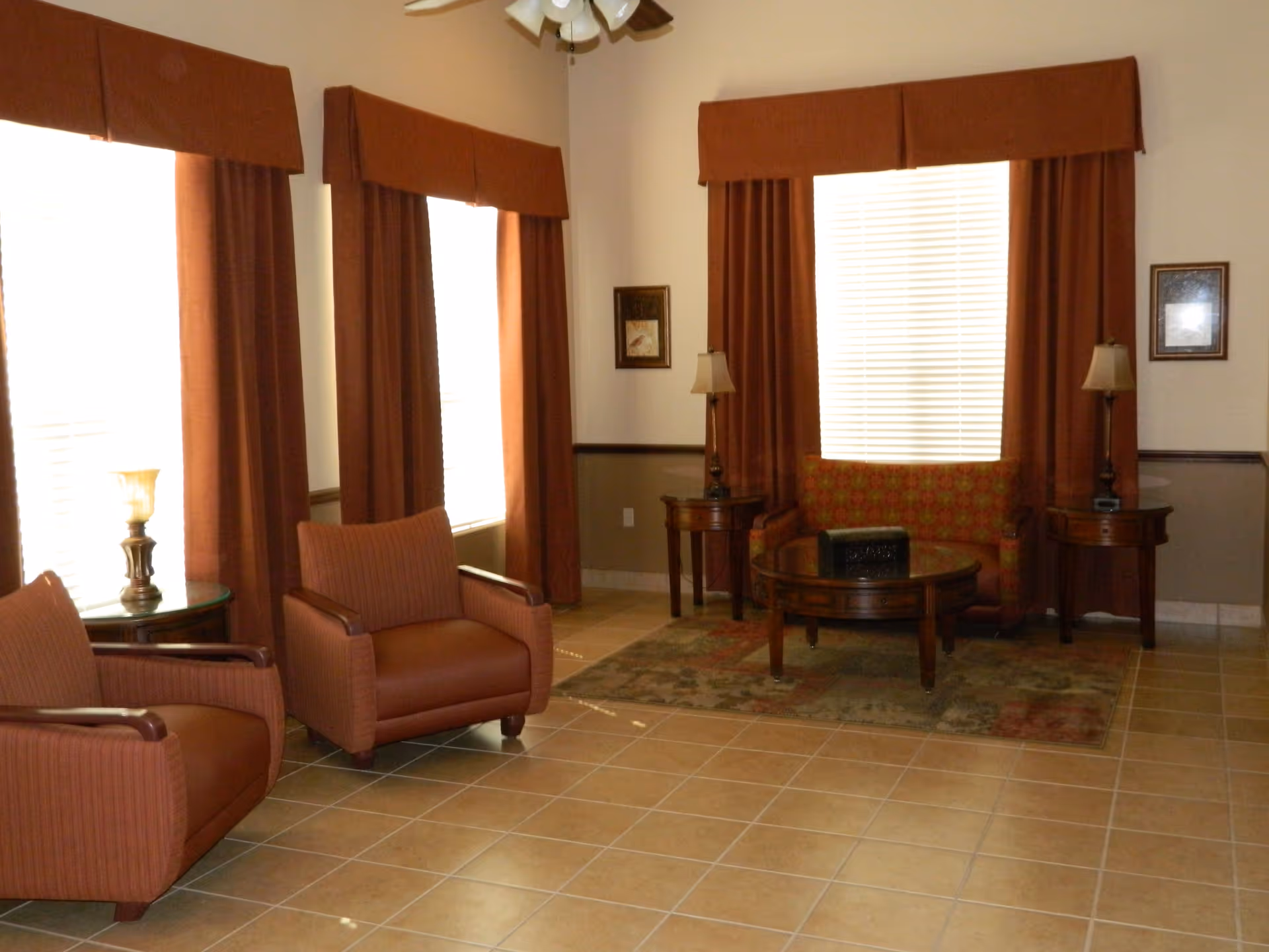 A cozy living room area with three brown upholstered armchairs and a patterned loveseat. The room has large windows with brown curtains and valances, two side tables with lamps, a round wooden coffee table on a patterned rug, and tiled flooring.