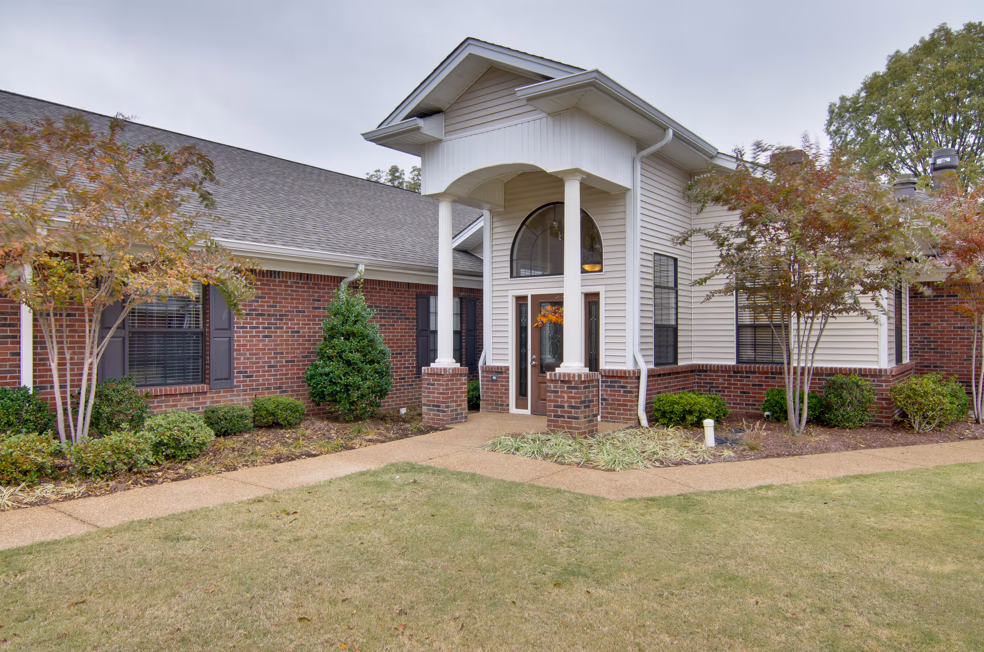 Front exterior view of Olive Grove Terrace Senior Living facility showing a brick and siding building with a covered entrance supported by white columns, surrounded by landscaped bushes and small trees.