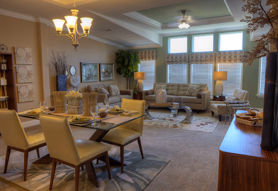 A well-lit living and dining area in a senior living facility with a glass dining table set for four, beige cushioned chairs, a beige sofa set with decorative pillows, two table lamps, large windows with blinds and valances, a ceiling fan, and various decorative items including plants and wall art.