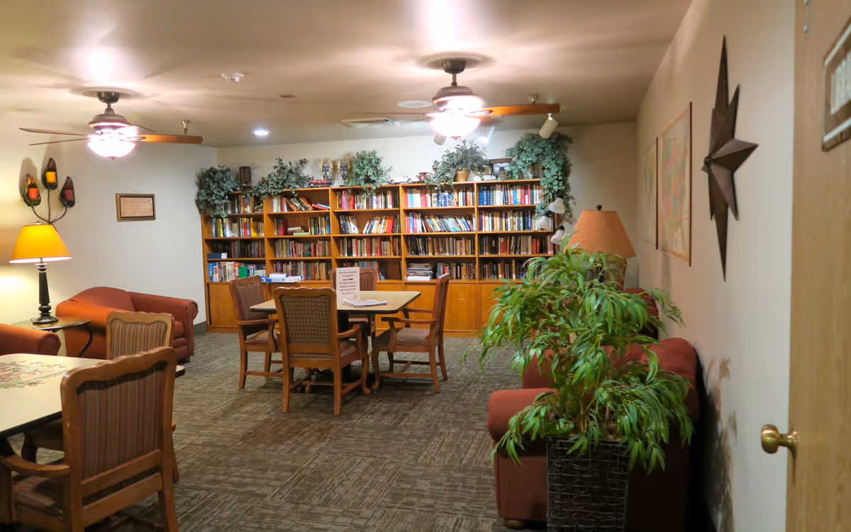 Cozy communal living area with bookshelves, a table and chairs, sofas, lamps, and plants.