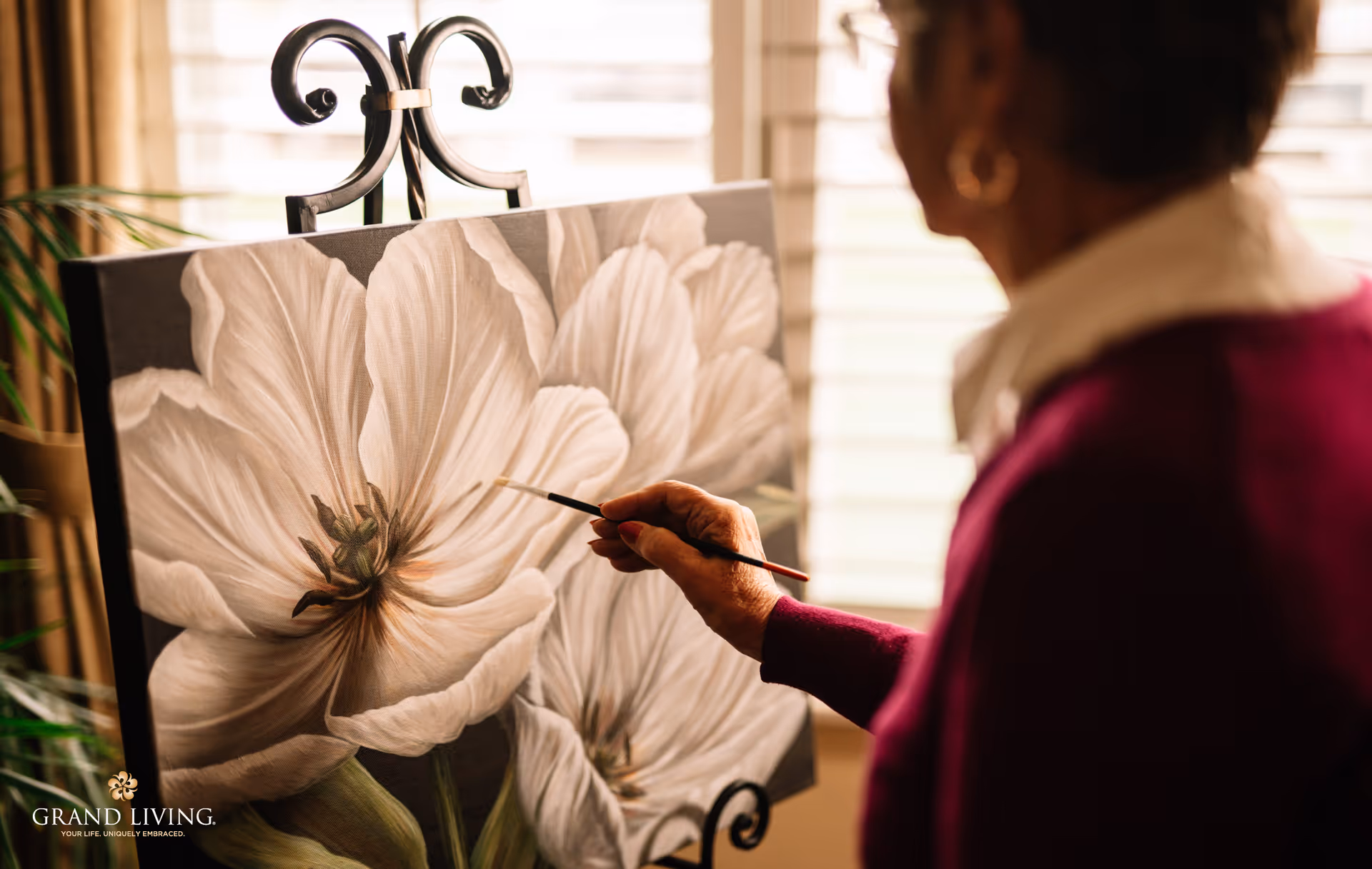 An elderly person painting a large canvas featuring white flowers indoors near a window with blinds. The person is holding a paintbrush and wearing a maroon sweater and white collared shirt.