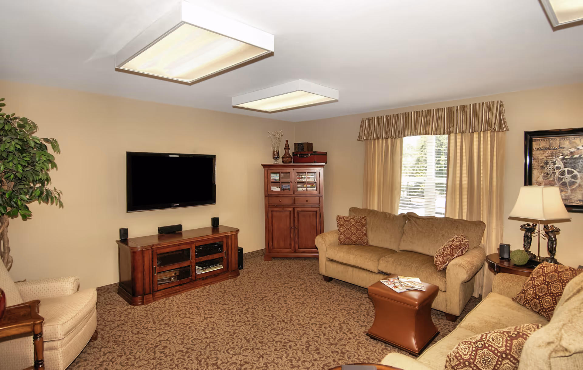 A cozy living room with beige walls and carpet, featuring a wall-mounted flat-screen TV above a wooden media console. There are two beige sofas with patterned cushions, a small brown ottoman, a wooden cabinet, a side table with a lamp, and a large window with beige curtains and a striped valance. A potted plant is visible on the left side.
