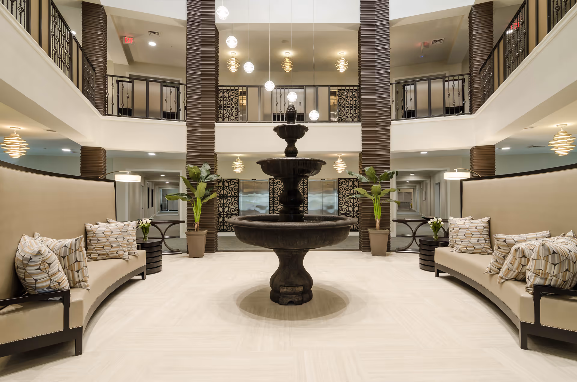 Spacious and elegant lobby area with a large black tiered fountain in the center, flanked by two curved beige sofas with patterned cushions. The space features tall pillars, potted plants, hanging pendant lights, and a second-floor balcony with decorative railings.