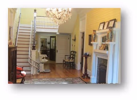 Interior view of a home featuring a wooden staircase with a stairlift, a chandelier hanging from the ceiling, a fireplace with decorative items on the mantel, wooden flooring, and chairs placed along the walls.