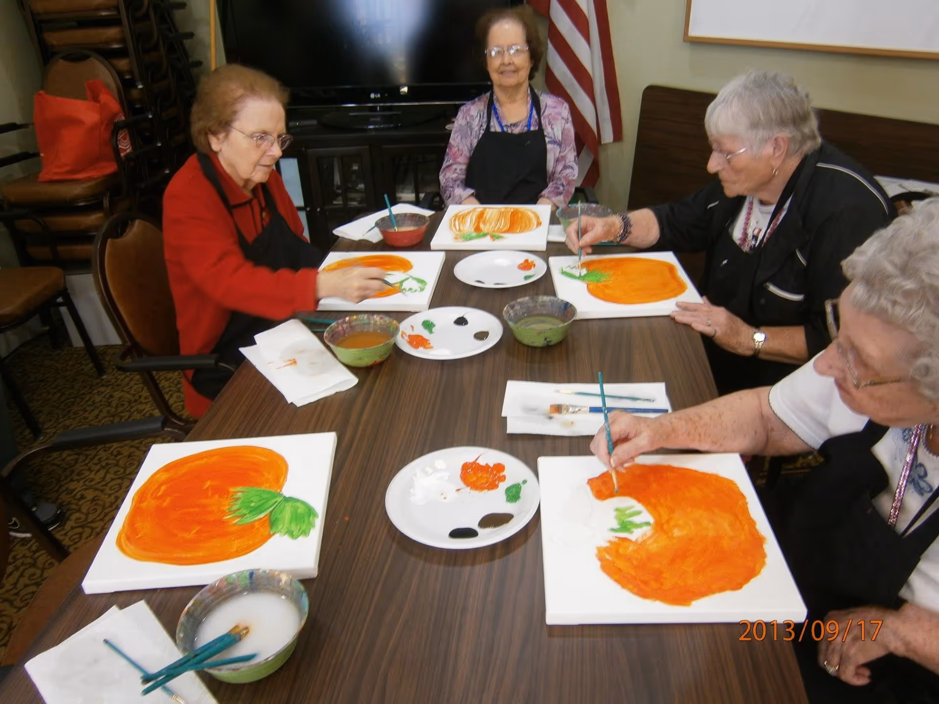 Four elderly women sitting around a table painting large orange pumpkins on canvases in a communal room with an American flag and a television in the background.