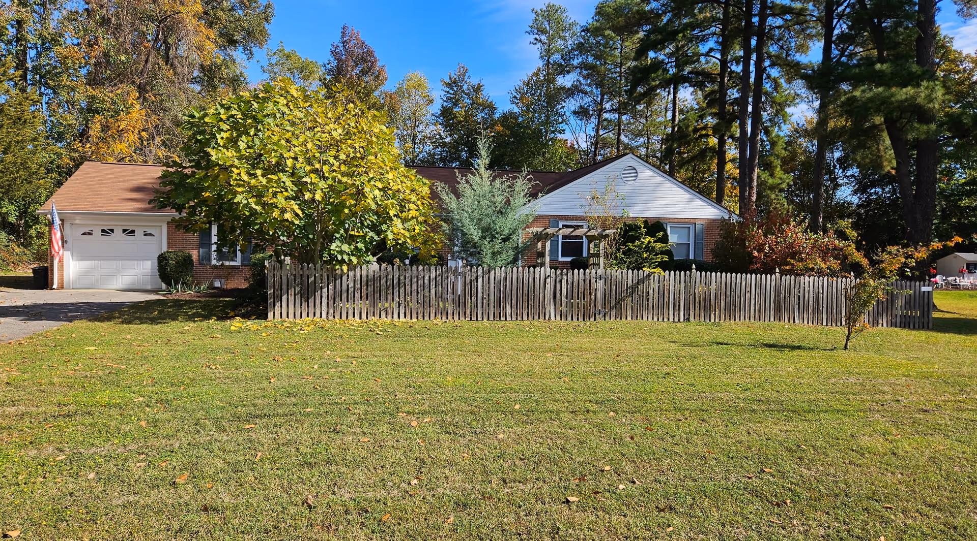 Single-story brick and white siding house with a brown roof, surrounded by a wooden picket fence and various trees, with a large grassy lawn in the foreground under a clear blue sky.