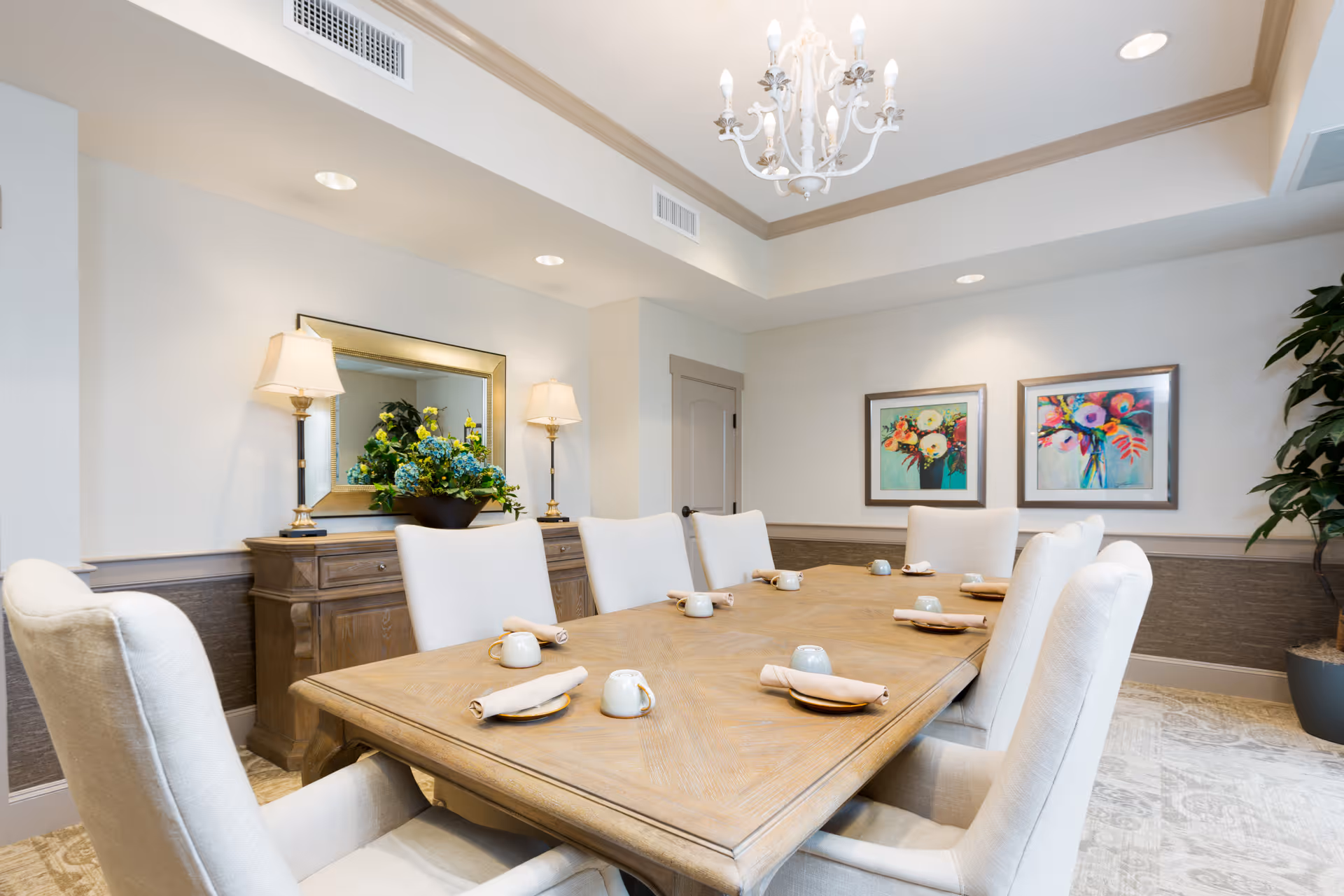 A well-lit dining room with a large wooden table set for eight, featuring beige upholstered chairs, rolled napkins on plates, and small cups. The room has a chandelier hanging from a tray ceiling, two framed floral paintings on the wall, a wooden sideboard with a large mirror and two lamps, and a potted plant in the corner.