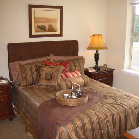 A neatly made bed with striped brown and beige bedding, multiple decorative pillows, and a round woven tray with items on it. The bed has a dark brown headboard. On either side of the bed are wooden nightstands with drawers, one holding a lamp with a beige shade and a small clock. A framed picture hangs on the wall above the bed, and a window is visible to the right letting in natural light.