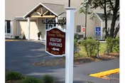 Outdoor view of a parking area at Welles Country Village with a sign indicating visitor parking. The sign is mounted on a white post and the building entrance is visible in the background along with some greenery and a handicapped parking sign.