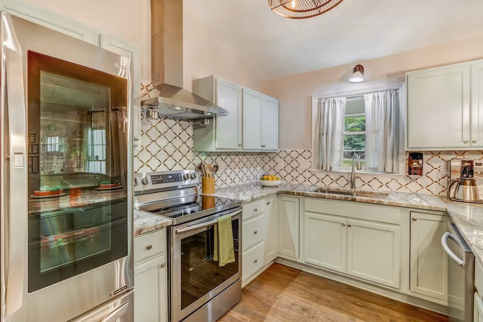 A modern kitchen with stainless steel appliances including a refrigerator and stove. The kitchen features white cabinets, a patterned tile backsplash, granite countertops, a window with checkered curtains, and wooden flooring.