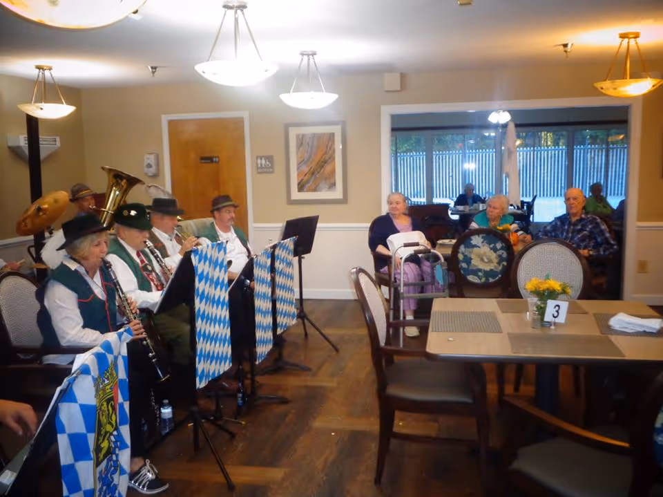 A small band dressed in traditional attire is playing musical instruments in a dining room area of a senior living facility. Several elderly residents are seated at tables and watching the performance. The room has wooden floors, beige walls, and ceiling lights. There are placemats and a small flower arrangement on the tables.