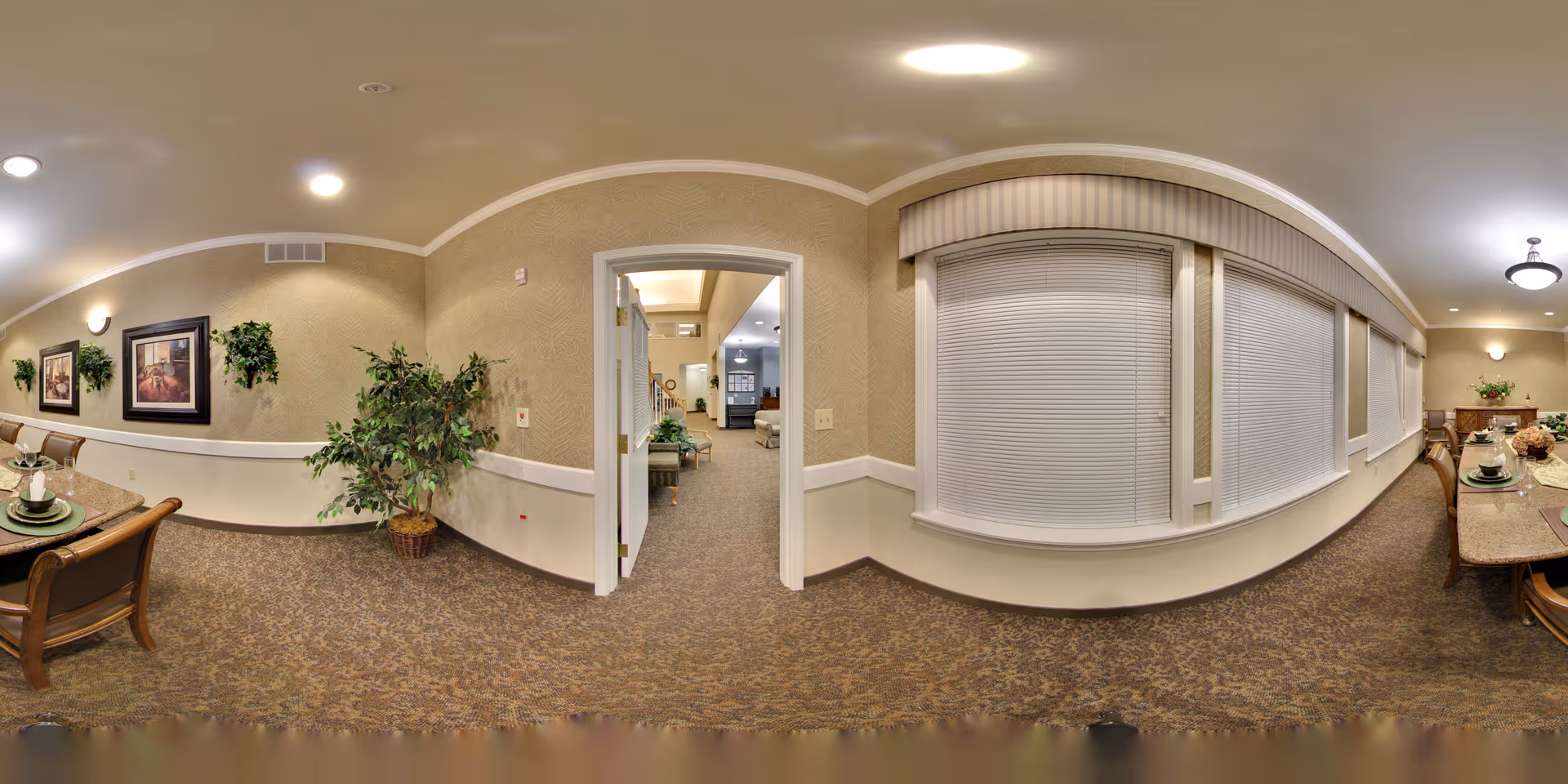 A panoramic view of a senior living facility interior showing a dining area with a long table set with plates and cups on the right, beige walls with framed artwork and potted plants, carpeted floor, and a doorway leading to a hallway with seating and additional rooms visible in the background.