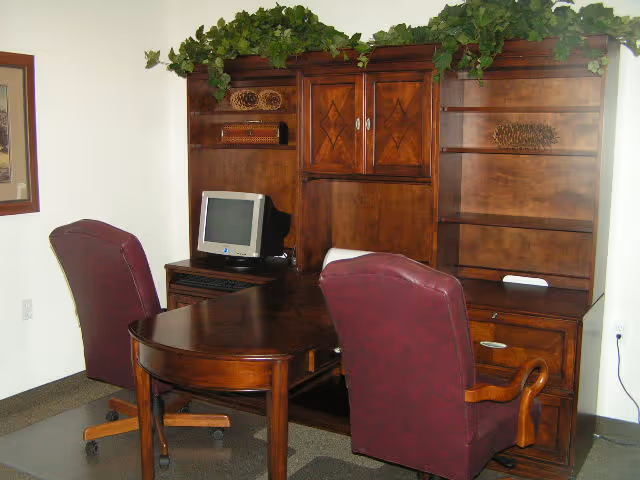 Office area with a dark wooden desk and matching hutch, two maroon leather swivel chairs, a small CRT computer monitor, and decorative greenery on top of the hutch.