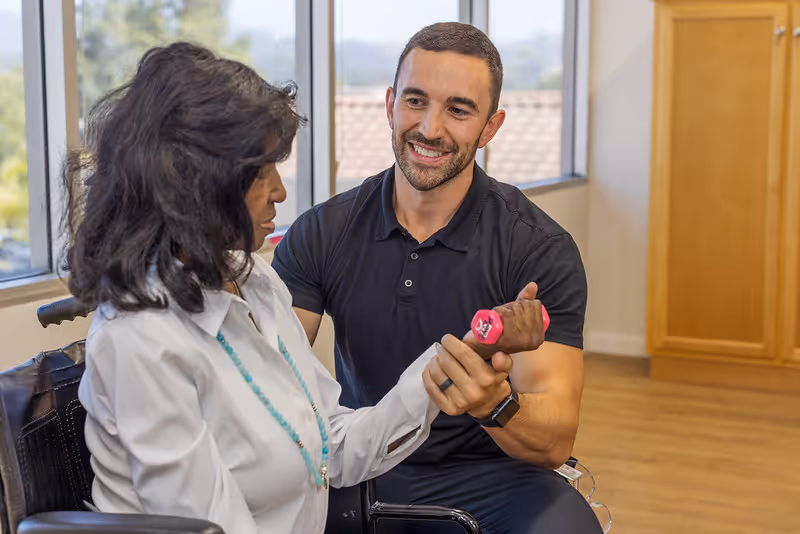 A man in a black polo shirt assists a woman in a wheelchair with lifting a small pink dumbbell in a room with large windows and wooden cabinets.