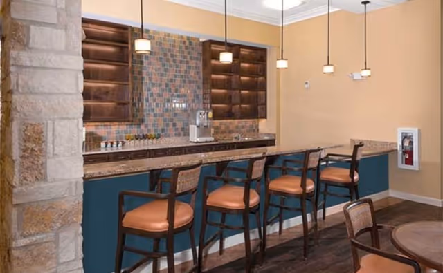 Community bar area with a tiled backsplash, granite countertop and four high chairs under pendant lights.