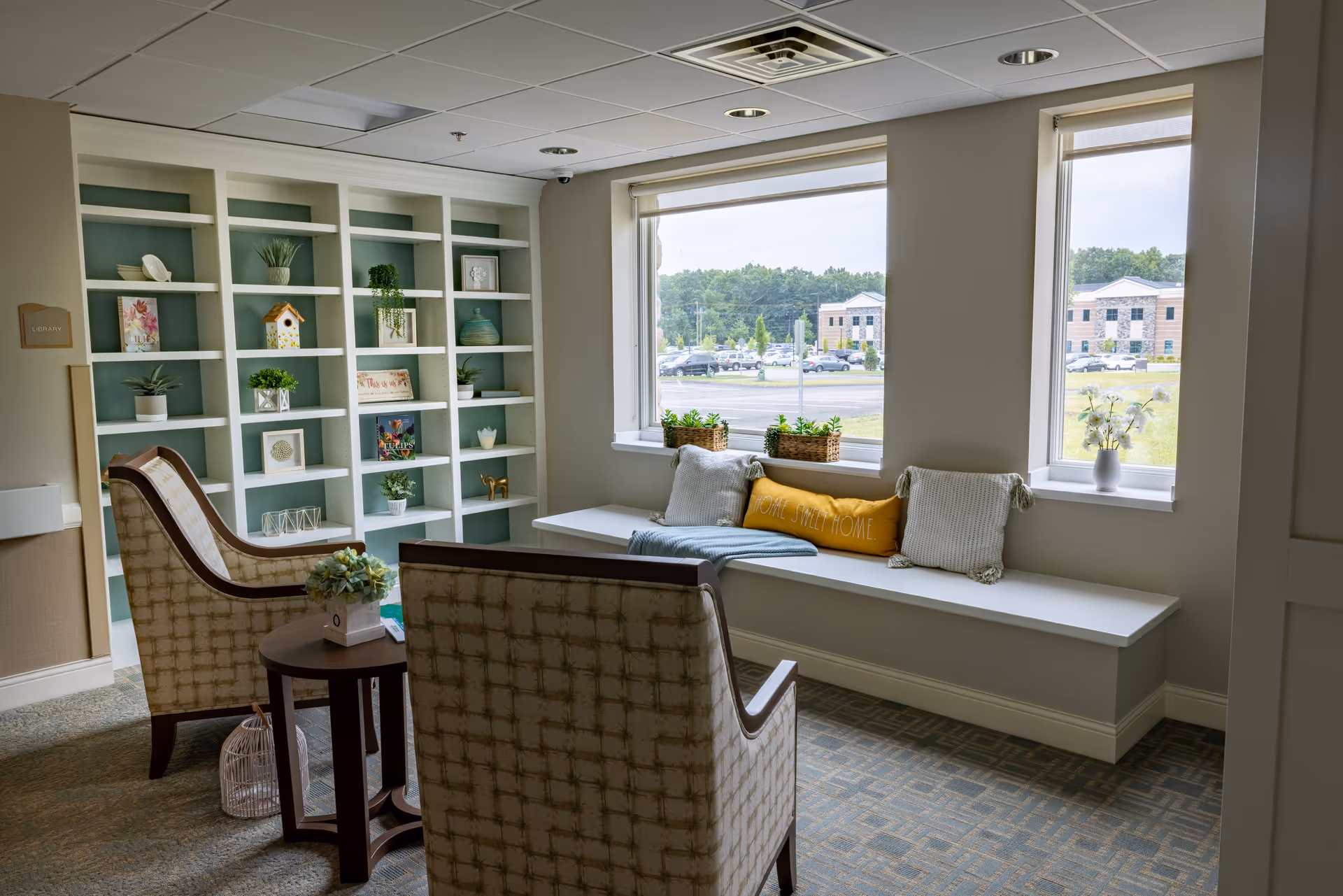 A cozy seating area in a memory care assisted living facility with two upholstered armchairs facing a small round wooden table. Behind the chairs is a built-in white bookshelf decorated with plants, books, and decorative items. A window seat with cushions and pillows, including one that says 'Home Sweet Home,' is positioned under two large windows overlooking a parking lot and trees outside.