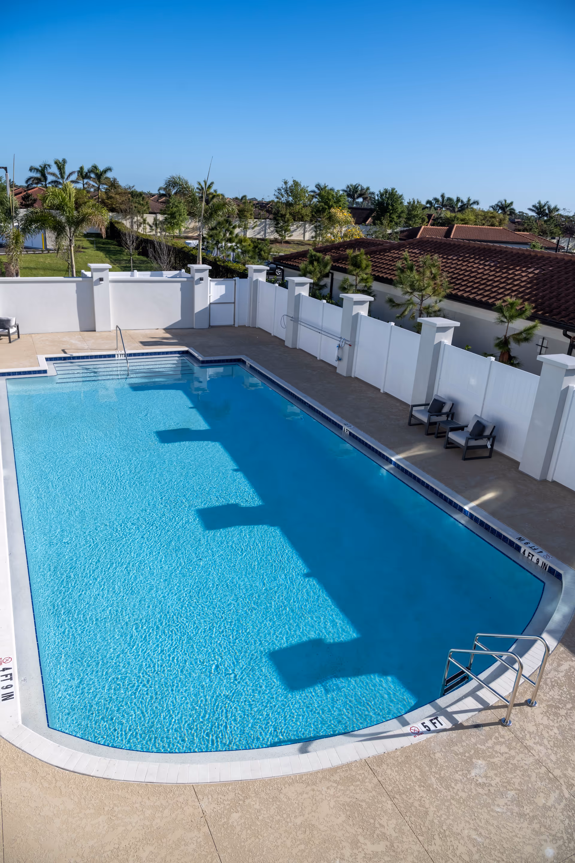 Outdoor swimming pool with clear blue water surrounded by a beige concrete deck. There are two chairs with a small table on the right side near a white fence. Palm trees and other greenery are visible beyond the fence under a clear blue sky.