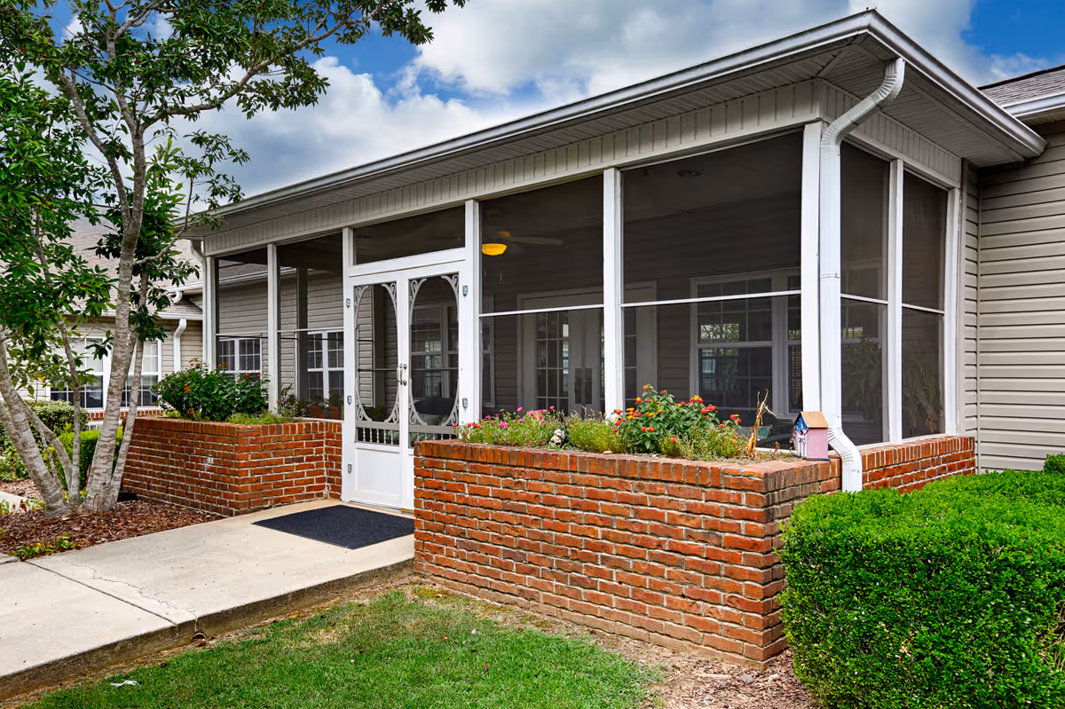 Exterior view of a screened porch attached to a building with beige siding. The porch is surrounded by a low red brick wall with flower beds on top. There is a white screen door in the center, a concrete walkway leading to the door, and green bushes and a tree nearby under a partly cloudy sky.