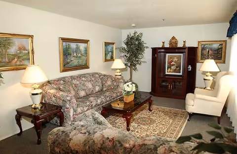 A cozy living room in a senior care facility with floral sofas, lamps, and a cabinet.