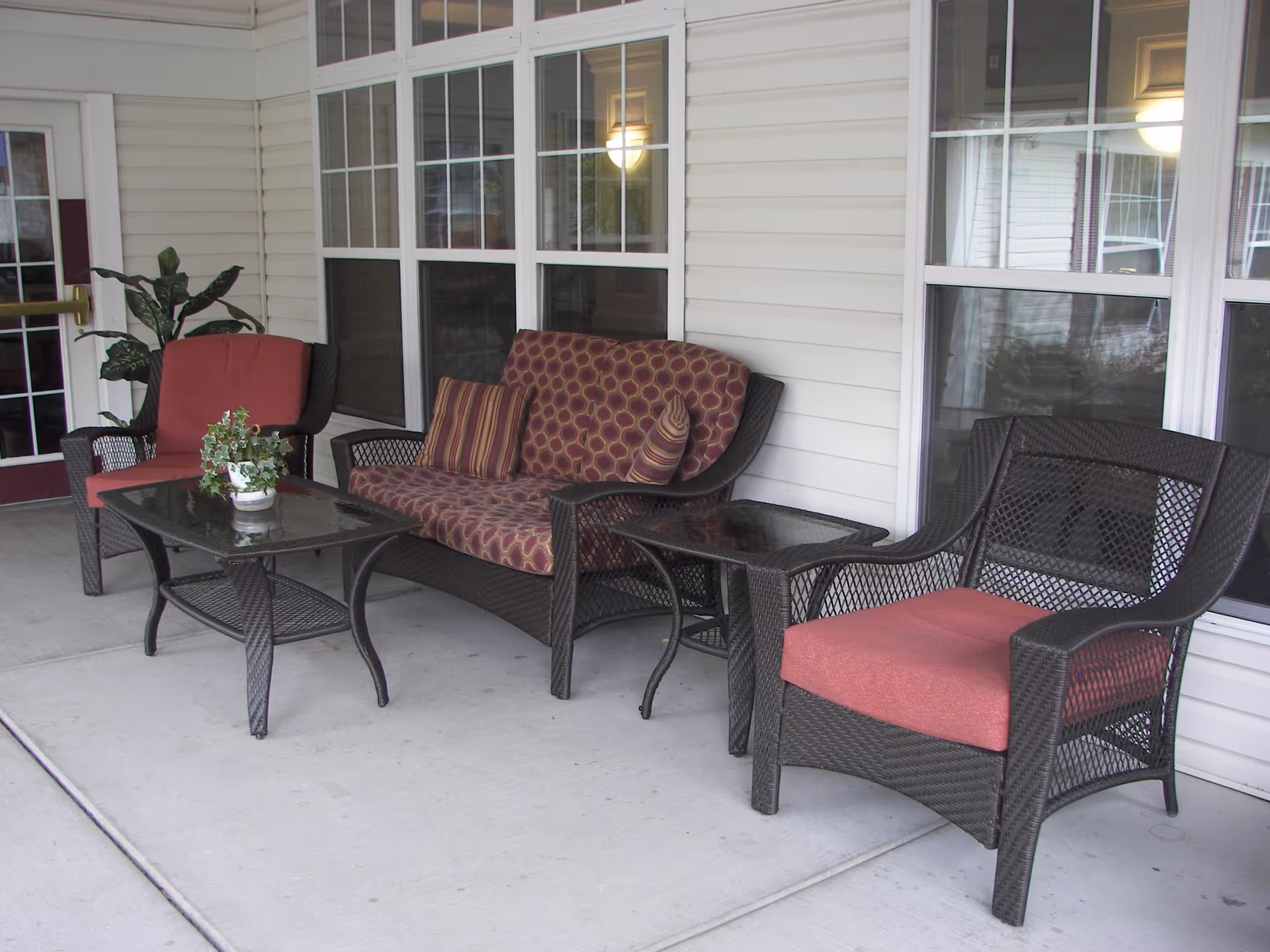 Outdoor seating area with dark wicker furniture including two armchairs with red cushions, a loveseat with patterned cushions, a glass-top coffee table with a potted plant, and a side table, all set on a concrete patio in front of windows and a door.